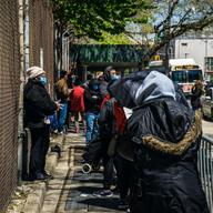 people of color wearing face masks line up outside of a building along a wire fence