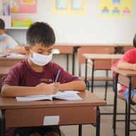 a young asian boy with a face mask writes in a notebook in a classroom. two other students are in the class seated several desks away