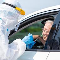 an older man in a white car leans slightly out the window to receive a swab up his nose from a person in a haz-mat suit