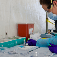 a nurse in a makeshift tent wearing protective gear readies multiple test kits in clear plastic containers