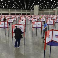 Voting stations with U.S. flags on them are set up in a large hall. A few people stand at different stations.