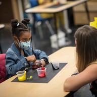 A little girl mixes paint in a classroom wearing a mask.