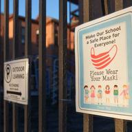 A school fence with a sign with a mask and cartoon children on it that says 'Make our school a safe place for everyone! Please wear your mask!'
