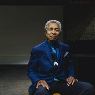 Musician Beverly Glenn-Copeland sits in a chair facing the camera. He's an older Black man with a short gray Afro, and wears a blue suit, dress shirt, and tie.