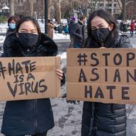 two asian women wearing masks and jackets outside in the snow carrying cardboard signs that say 'hate is a virus' and 'stop asian hate'