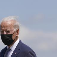 Joe Biden wears a black face mask and a navy suit. He looks into the distance, with blue sky in the background.