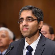 an indian man wearing glasses and a suit tie with a small microphone in front of him, with a small crowd of people behind him