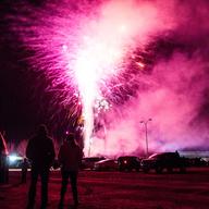Three people stand in a parking lot while large pink fireworks explode in front of them.