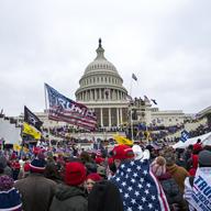 In the background is the U.S. capitol building on the day of the January 6th insurrection, with crowds visible on its steps. In the foreground are more people packed together waving Trump flags.