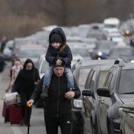 A man in the center of the frame with a child riding his shoulders leading a scant line of other blurred travelers on the side of the road next to countless cars packed bumper to bumper on the road.