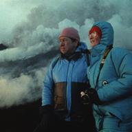 Katia (right) and Maurice Krafft (left), in blue winter jackets, gaze upon a volcano in the distance as smoke, steam and ash swirl behind them. 