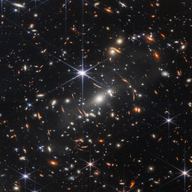 In this image, captured by JWST, has a background of space is black. Thousands of galaxies appear all across the view. Their shapes and colors vary. Some are various shades of orange, others are white