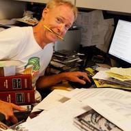 John Schaefer with a pencil in his mouth sitting at a very messy desk
