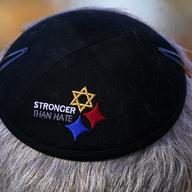 A black kippah on a man's head is shown with the words 'stronger than hate' embroidered in white, next to a golden Star of David and two diamonds, one in blue and one in red. 