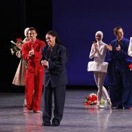 A smartly dressed woman onstage, surrounded by colorfully costumed dancers.