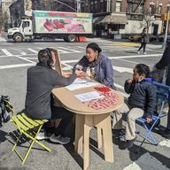 A reporter interviewing a local resident as they sit across from a table on Amsterdam Avenue and West 107th Street in Manhattan.