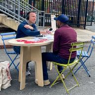 A reporter is interviewing a man across a table on a street in Staten Island. A sign on the table reads 'share your story with WNYC.'  Buttons and pens strewn the table. 