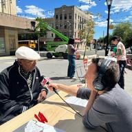 A reporter with a microphone interviewing a local resident across a table at Zion Triangle in Brownsville, Brooklyn. 