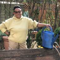 Community gardener Ena K. McPherson holding a blue-colored garden watering container. Shes' wearing a yellow sweatshirt and hat.