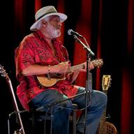 A man in a hat and a bright red shirt singing and strumming a small stringed instrument