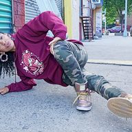 B-girl Ana 'Rokafella' Garcia is doing a break dance move on the street. She is wearing a purple sweatshirt and camouflage pants.