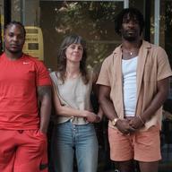 A woman wearing a plain white t-shirt stands between two men in front of the window of a store. One man is wearing a plain red shirt and red shorts and the other is wearing pink shorts, a plain white 