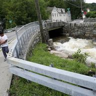 Pedestrians pass a local river whose heavy current flooded nearby Main Street, Monday, July 10, 2023, in Highland Falls, N.Y. Heavy rain has washed out roads and forced evacuations in the Northeast as