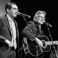 A singer, standing, and a seated guitarist performing onstage