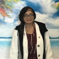 Marie Spivey is wearing glasses and smiling as she stands in front of a Caribbean-themed backdrop. She has a rainbow-colored pin on her sweater to celebrate Pride.  