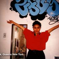 A young woman with a red blouse has her hands outstretched above her head to show off a graffiti tag on a wall behind her. 