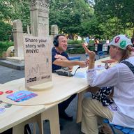 A reporter is interviewing a man across a table in Washington Square Park. A sign on the table reads 'share your story with WNYC.'  Buttons and pens strewn the table. 