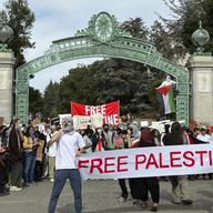 Two students hold a banner that says 'Free Palestine' in front of a green mental archway, with more people gathered in the background