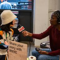 A reporter is interviewing a woman across a table at Industry City in Sunset Park, Brooklyn. A sign on the table reads 'share your story with WNYC.'  Buttons and pens strewn the table. 