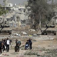 A group of eight men walk along a path littered with debris. In the background two tanks have their guns pointed at the group