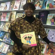 Masani Barnwell George is standing in front of shelves full of books in her story called Source of Knowledge. She has a big smile and is holding a children's book titled 'Love is Me'