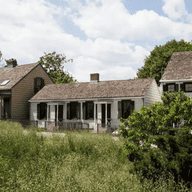 A set of three house( known as the Historic Hunterfly Road Houses) sit between a green field in the forefront and a set of trees and blue sky with white fluffy clouds in the back framing the photo.