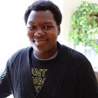 Emmanuel stands by window with sun beaming on the right of his face, that has a slight smile on it. He's wearing a button up cardigan over a black shirt with the words ; 'ANT', 'AN', & 'K' visible.