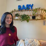 A woman wearing a red sweater stands in front of a sign saying 'baby.'