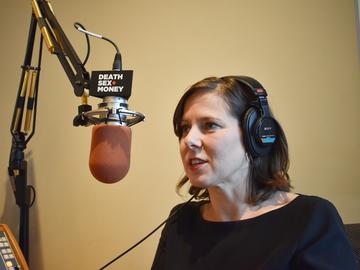 Anna Sale is wearing headphones, talking into a microphone in a studio. She has short wavy hair and is wearing a black top. She is mid-conversation.