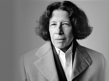 A black and white photo of writer Fran Lebowitz from the shoulders up. She’s an older white woman with dark hair wearing a peacoat, a blazer, and a white button up with the top button open.