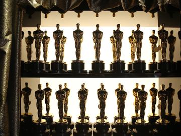 A close-up photograph of a two-tiered shelf displaying dozens of Oscar awards lined up in pairs, framed by a shiny gold curtain and backlit from a display light behind the shelf.