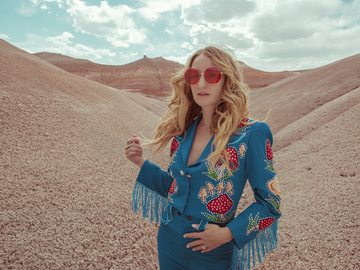 A photographed portrait of country singer, Margo Price, standing in a sandy desert scene, wearing big, red octagonal sunglasses, and a western-style blue suit with embroidered mushrooms. 