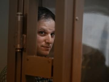 A photograph of detained Wall Street Journal reporter, Evan Gershkovich, looking toward the camera, with his face obscured by the metal edges of the door containing him.