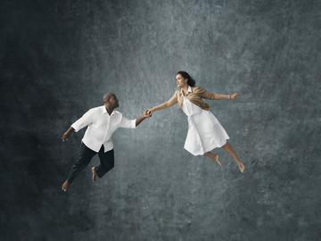 Two performers, a man and a woman, suspended in midair touching hands