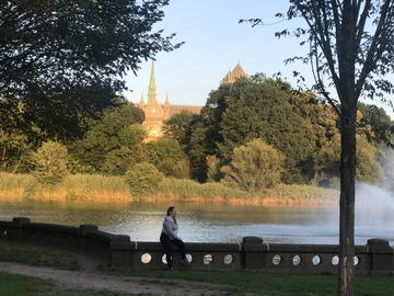 A woman is sitting on a stone wall in front of a lake as the sun sets in Essex County Branch Brook Park. There are a lot of leafy trees around and a cathedral in the far background. 