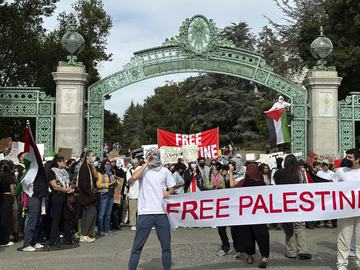 Two students hold a banner that says 'Free Palestine' in front of a green mental archway, with more people gathered in the background