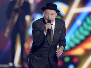 A photograph of salsa singer Ruben Blades, singing into a wireless microphone, wearing a dark blue suit, green tie and pocket square, and black pork pie hat. 