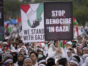 Two signs are held above a crowd of protesters. One says 'We Stand With Gaza.' The other says 'Stop Genocide in Gaza.'