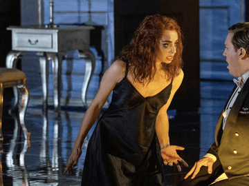 A dramatic scene from a stage performance, featuring a woman with curly red hair in a black dress and a man in formal attire. The woman appears distressed, yelling at a man in a suit
