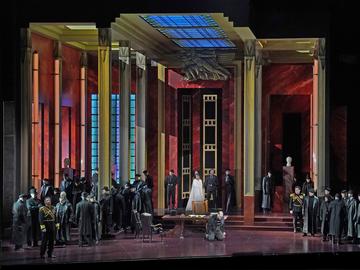 An opera stage set in a grand and opulent interior featuring tall columns, stained glass windows, and red marble walls. The scene includes a large group of performers in formal, dark attire, some wear